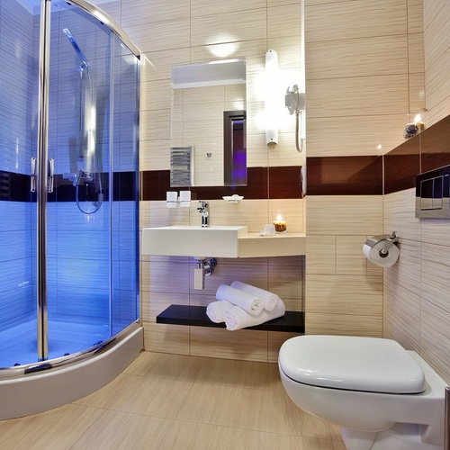 Hotelowa łazienka z niebiesko podświetloną kabiną prysznicową, podwieszaną umywalką, ręcznikami i ścienną toaletą.