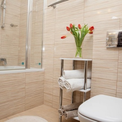 Łazienka z wanną i prysznicem za szklaną ścianką, beżowe płytki, chromowana armatura, półka z ręcznikami i tulipanami.