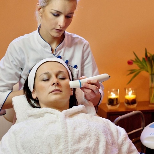 Profesjonalny zabieg spa w pokoju zabiegowym: kosmetyczka wykonuje zabieg urządzeniem na twarzy, świece i tulipany w tle.