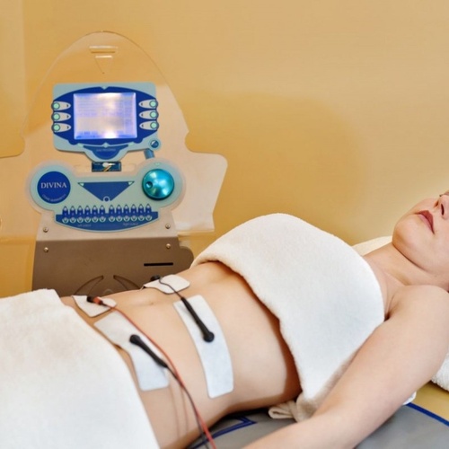 Kobieta na zabiegu spa, leżąca na łóżku z ręcznikiem; elektrody na brzuchu pod nowoczesnym aparatem do modelowania sylwetki, relaksująca atmosfera