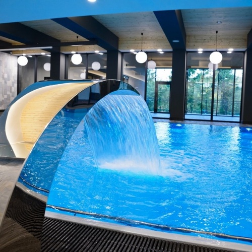 Hotelowy basen wewnętrzny z metalową kaskadą wodną, dużymi oknami z widokiem na zieleń i kulistymi lampami.