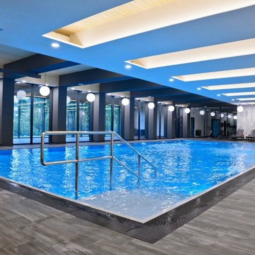 Nowoczesny kryty basen hotelowy z niebieską wodą, stalowymi poręczami, przeszklonymi ścianami, okrągłymi lampami i leżakami