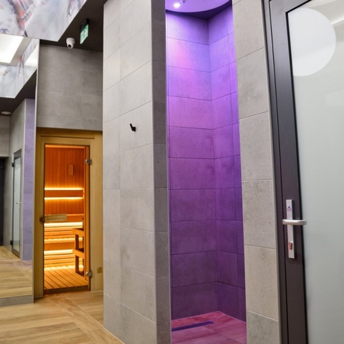Strefa spa z otwartą drewnianą sauną o ciepłym świetle, fioletowo podświetloną kabiną i nowoczesnymi płytkami.