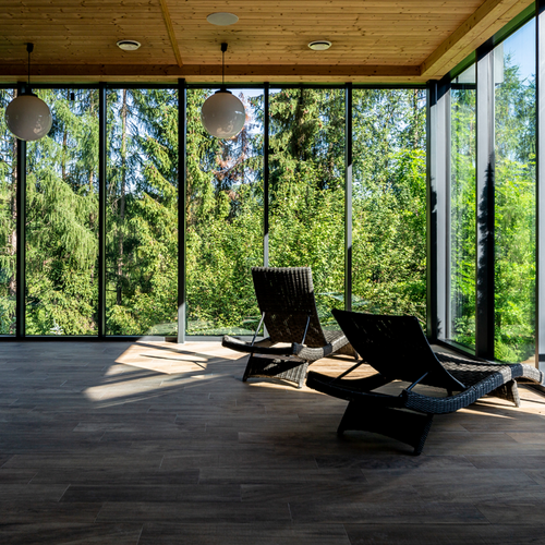 Pokój relaksu spa z przeszklonymi ścianami, wiklinowymi leżakami, drewnianym sufitem i panoramicznym widokiem na las.