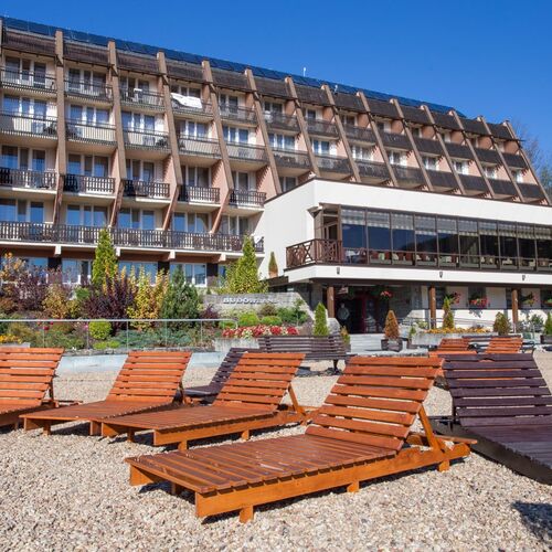 Hotel z tarasem i przeszkloną restauracją, rzędem drewnianych leżaków na żwirowej plaży, balkonami pokoi i zadbanym ogrodem