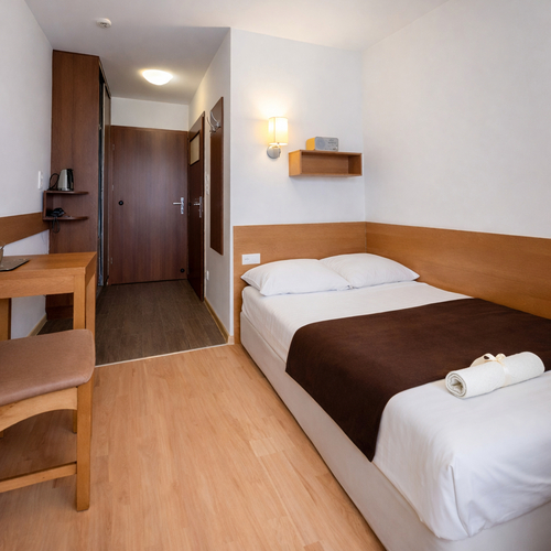 Przytulny, kompaktowy pokój hotelowy z pojedynczym łóżkiem, biurkiem, czajnikiem, półką z radiem, lampą ścienną i zwiniętym ręcznikiem.