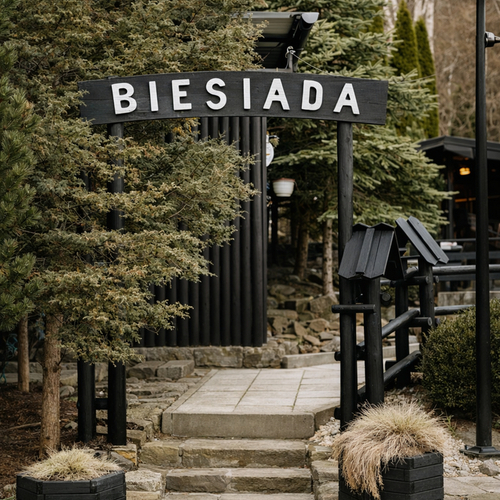 Wejście do rustykalnej restauracji Biesiada: drewniana brama, kamienne schody, iglaste drzewa i donice