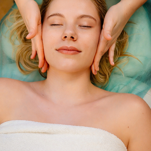 Kobieta z zamkniętymi oczami podczas relaksującego masażu twarzy w pokoju spa, na leżance z białym ręcznikiem