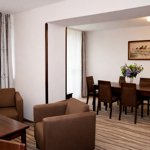 Przestronny apartament z salonem i jadalnią: fotele i stolik, drewniany stół na 6 osób z bukietem, jasne zasłony, miękki dywan i ciepłe oświetlenie.