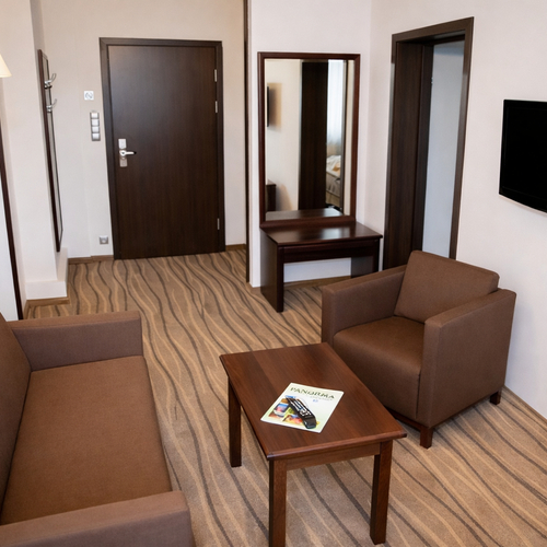 Przytulny kącik pokoju: brązowa sofa i fotel, drewniany stolik z magazynem, lustro i TV na ścianie, wejście do pokoju.