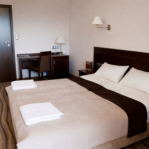 Hotelowy pokój z dużym łóżkiem, ciemnym drewnianym zagłówkiem, lampkami przy łóżku, biurkiem z krzesłem i złożonymi ręcznikami.