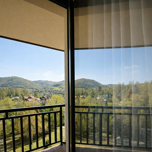 Balkon pokoju z przeszklonymi drzwiami i tiulem, czarną poręczą i panoramicznym widokiem na zielone wzgórza i domki.