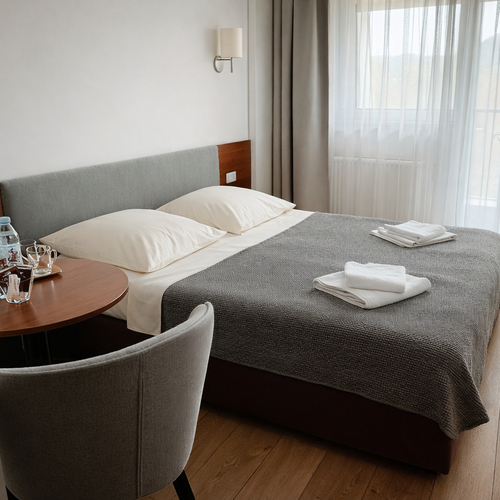 Dwuosobny pokój hotelowy z wygodnym łóżkiem, szarą narzutą, złożonymi ręcznikami, stolikiem z butelkami wody i jasnym oknem z balkonem.