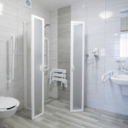 Przystosowana łazienka hotelowa z bezprogowym prysznicem z składanym siedziskiem, poręczami, umywalką i suszarką.