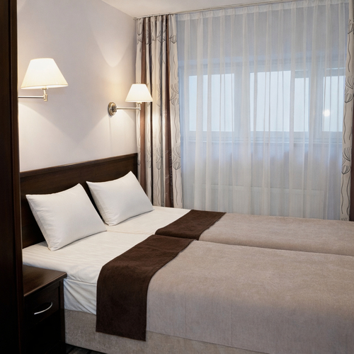 Pokój hotelowy z dwoma przystawionymi łóżkami, białą pościelą, brązowymi narzutami, ciemnymi meblami, lampami ściennymi i zasłonami.
