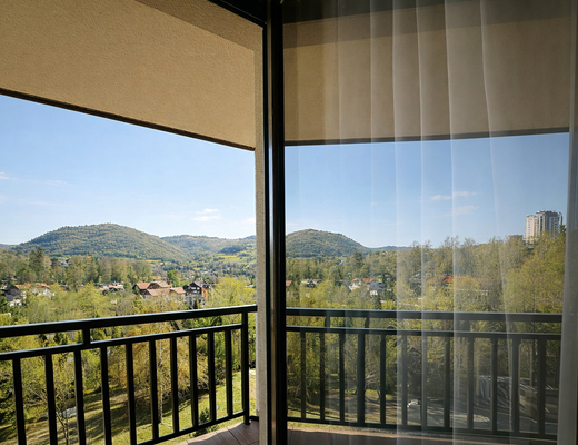 Balkon pokoju z przeszklonymi drzwiami i tiulem, czarną poręczą i panoramicznym widokiem na zielone wzgórza i domki.