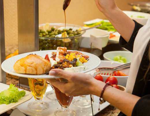 Gość polewa sosem sałatkę przy bufecie: talerz z bułką, serem, kukurydzą i warzywami, miska oliwek w tle