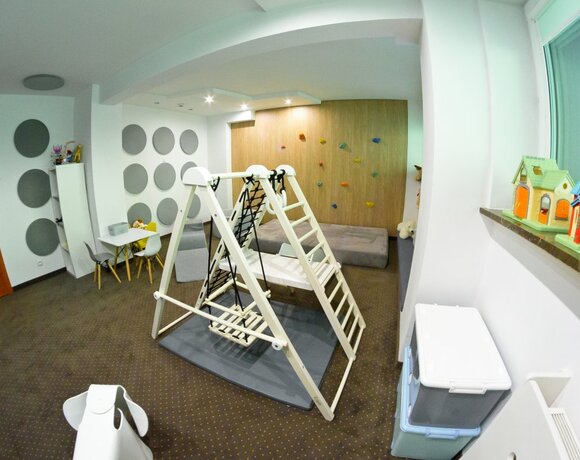 Sala zabaw hotelowa z drewnianą drabinką i huśtawką, miękkimi matami, ścianką wspinaczkową, stolikiem i zabawkami.
