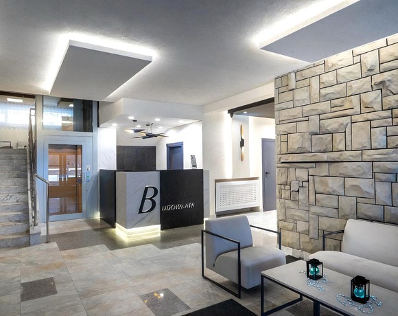 Eleganckie lobby z marmurowymi schodami i windą, podświetloną recepcją z logo B, kamienną ścianą i strefą wypoczynku.