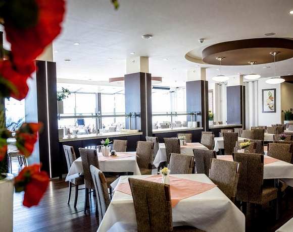 Przestronna restauracja hotelowa z białymi obrusami, łososiowymi serwetkami, stolikami, bufetem przy oknach i kwiatami.