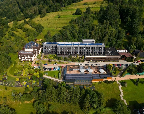 Widok z lotu ptaka na górski hotel z balkonami, tarasem spa z parasolami, panelami słonecznymi i parkingiem wśród lasu.