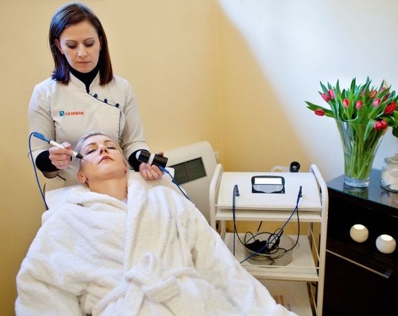 Kosmetyczka wykonuje zabieg na twarz urządzeniem, klientka w białym szlafroku na leżance, wazon z czerwonymi tulipanami.