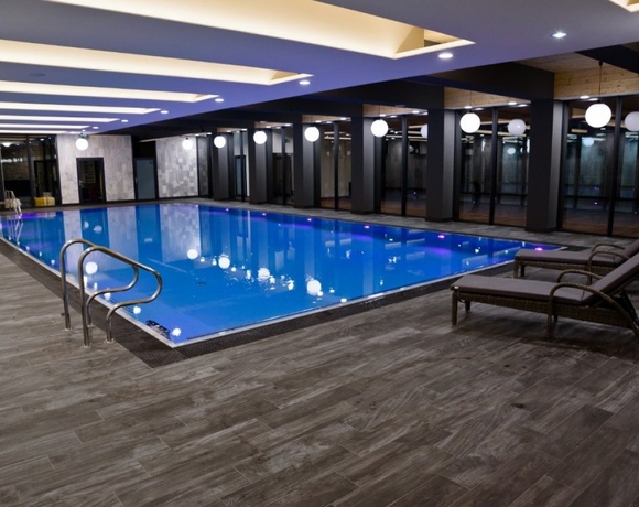 Wewnętrzny basen hotelowy z niebieskim podświetleniem, wygodnymi leżakami, stalową poręczą i dużymi oknami.