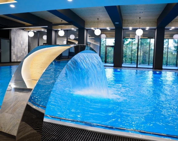Hotelowy basen wewnętrzny z metalową kaskadą wodną, dużymi oknami z widokiem na zieleń i kulistymi lampami.