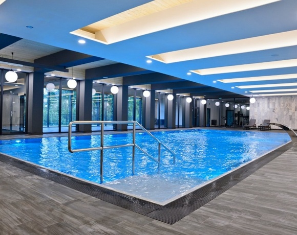 Nowoczesny kryty basen hotelowy z niebieską wodą, stalowymi poręczami, przeszklonymi ścianami, okrągłymi lampami i leżakami