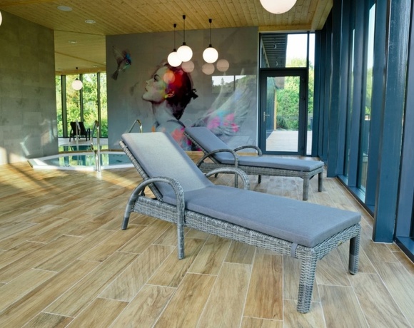 Strefa SPA z dwiema rattanowymi leżankami obok małego basenu, dużymi oknami, muralem i ciepłym drewnianym wykończeniem.