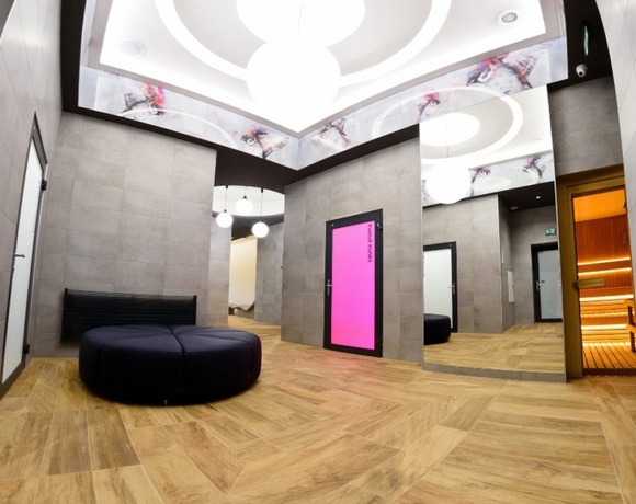 Nowoczesne spa - lobby z drewnianą podłogą, okrągłą czarną pufą, lustrem, różowo podświetlonymi drzwiami i widoczną sauną.