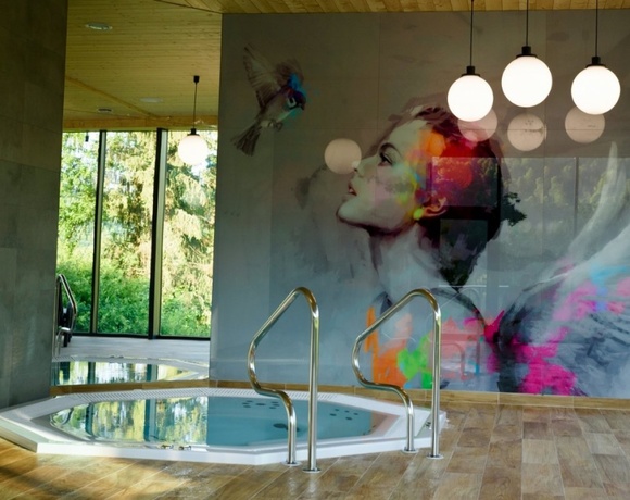 Wnętrze spa: jacuzzi z metalowymi poręczami, mural kobiety z kolorowymi skrzydłami, duże okna na zieleń.