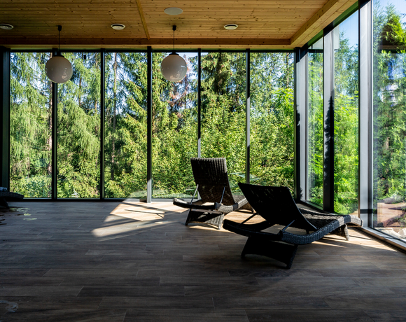 Pokój relaksu spa z przeszklonymi ścianami, wiklinowymi leżakami, drewnianym sufitem i panoramicznym widokiem na las.