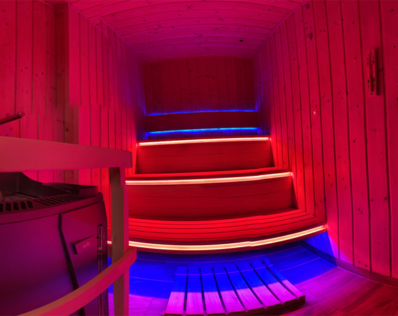 Drewniana sauna z piętrowymi ławkami, piecem do kamieni i nastrojowym czerwono-niebieskim oświetleniem LED