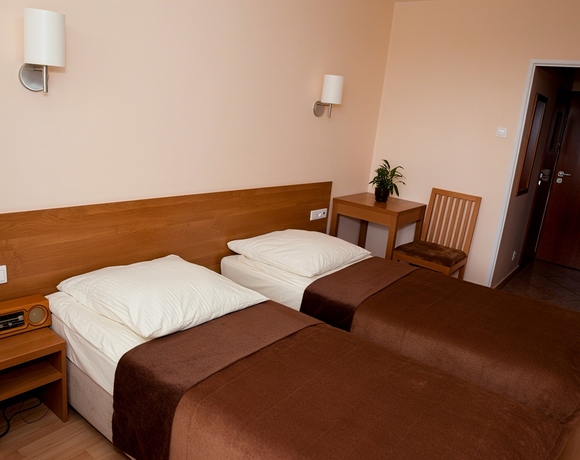 Dwa pojedyncze łóżka z drewnianym zagłówkiem i brązowymi narzutami, nocny stolik z radiem, lampy ścienne, biurko z rośliną.