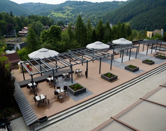 Taras hotelowy z drewnianym tarasem, pergolami i stolikami pod parasolami, donice i panoramiczny widok górski