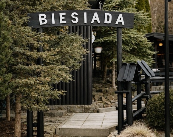 Wejście do rustykalnej restauracji Biesiada: drewniana brama, kamienne schody, iglaste drzewa i donice