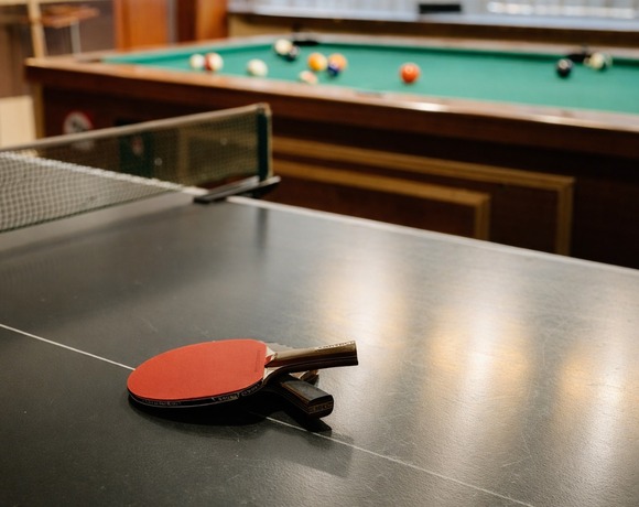 Stół do ping-ponga z dwiema rakietkami na środku, w tle stół bilardowy — przytulna sala gier hotelowych