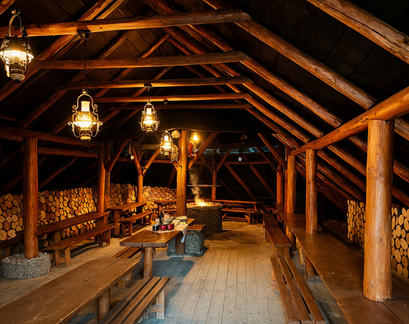 Rustykalna sala górskiej chaty: drewniane belki, długie stoły i ławy, centralne palenisko, stosy drewna i ciepłe lampy