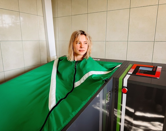 Kobieta relaksująca się w zielonej kapsule zabiegowej do odnowy z panelem sterowania, w jasnym, kafelkowanym pokoju spa