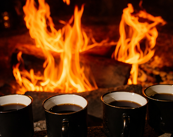 Cztery czarne kubki z gorącym napojem ustawione na kamieniu przed trzaskającym kominkiem, przytulna atmosfera