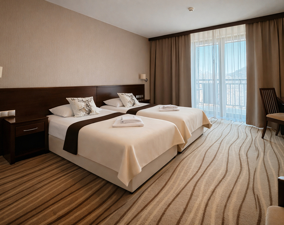 Pokój z dwoma pojedynczymi łóżkami, pościel, ozdobne poduszki, złożone ręczniki, ciemne meble, biurko i balkon z widokiem.