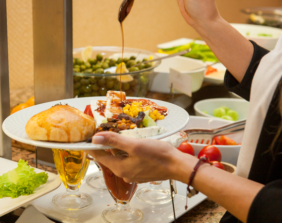 Gość polewa sosem sałatkę przy bufecie: talerz z bułką, serem, kukurydzą i warzywami, miska oliwek w tle