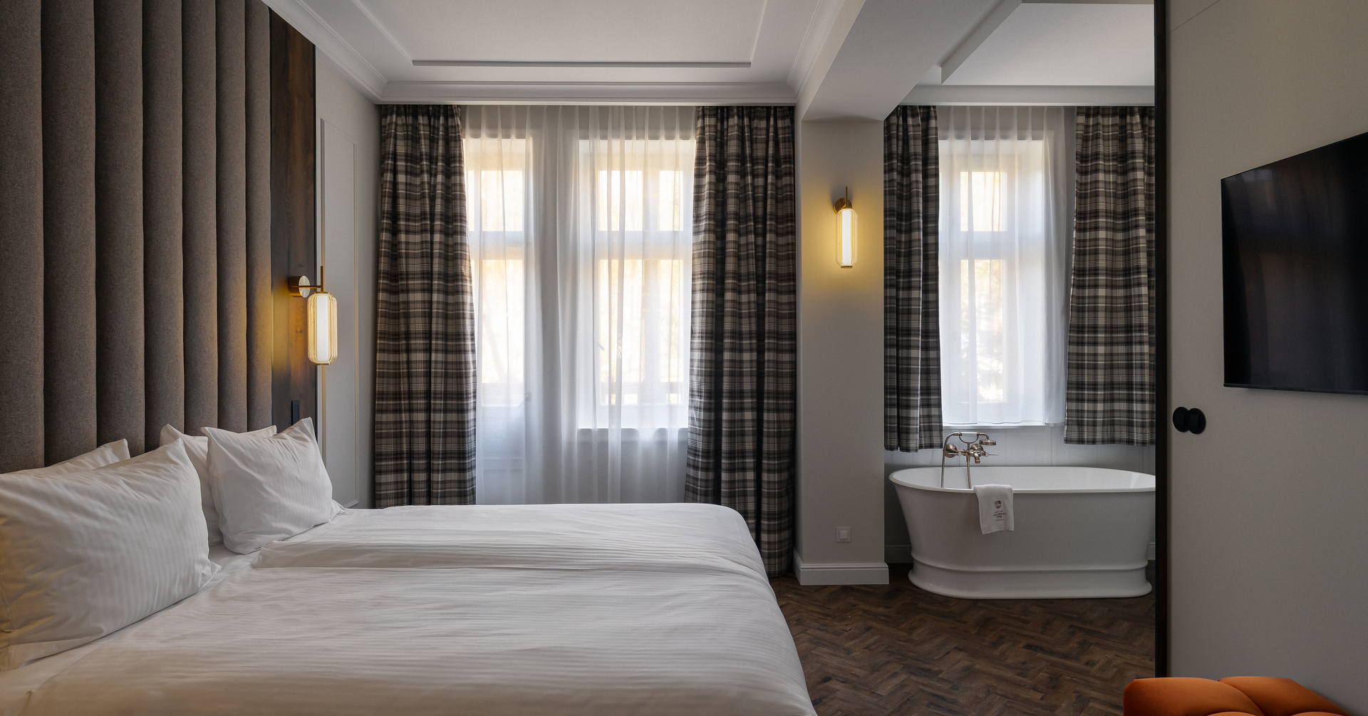 Łóżko king z wysokim tapicerowanym zagłówkiem, lampy ścienne, wolnostojąca wanna przy oknach z prześwitującymi zasłonami i drewnianą podłogą.