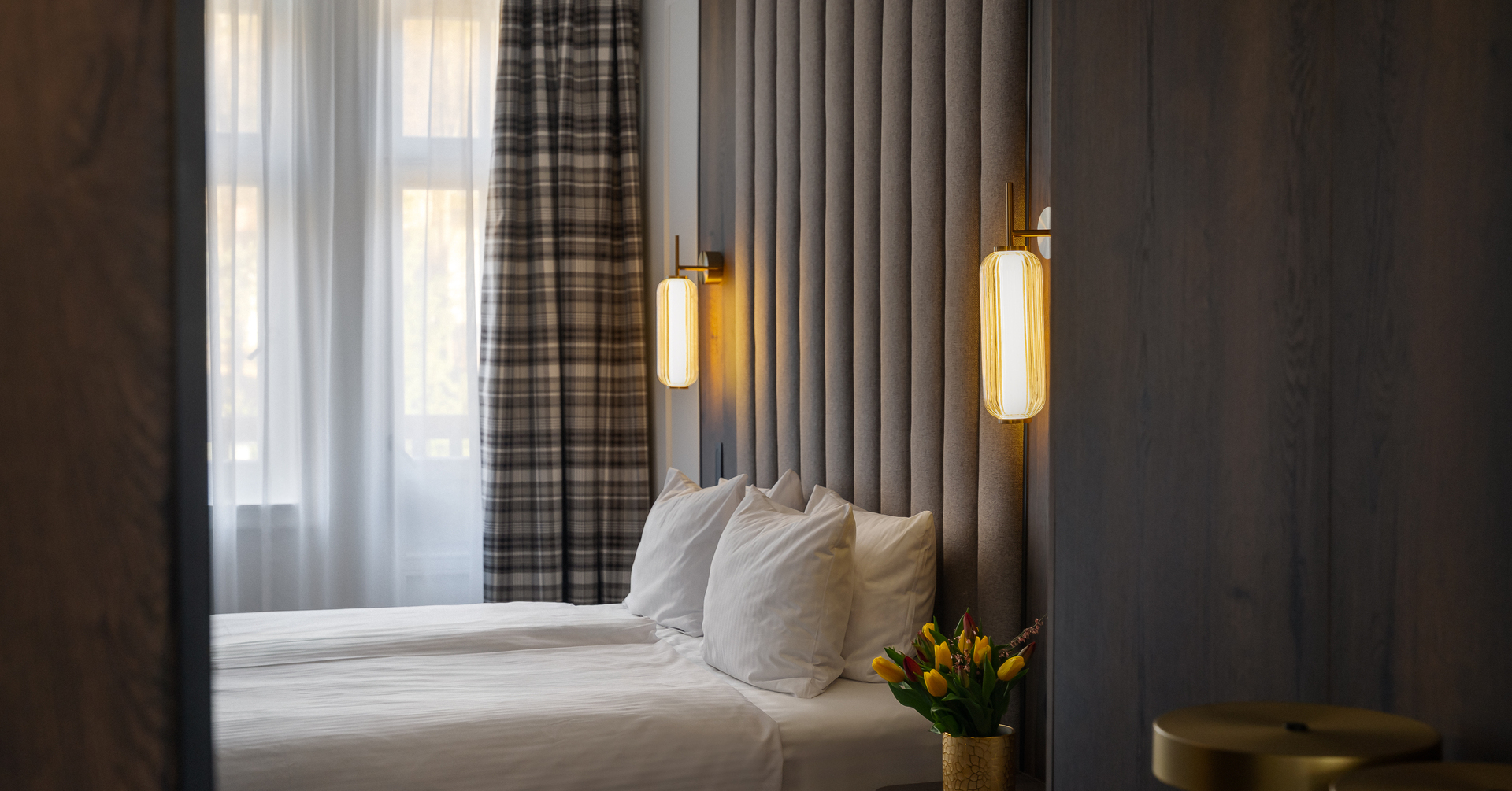 Nowoczesne dwa łóżka pojedyncze z wysokim tapicerowanym zagłówkiem, ciepłe kinkiety, firany, kratowane zasłony i bukiet tulipanów