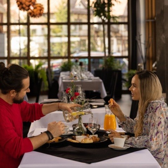 Pár užívající si snídani v hotelové restauraci u stolu s bílým ubrusem, croissanty a pomerančovým džusem, dřevěný kazetový strop a velká okna.