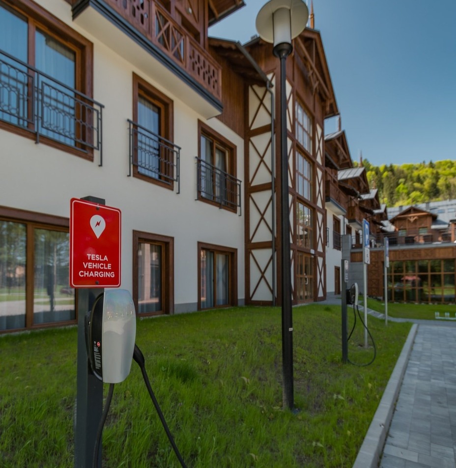 Stacja ładowania Tesla przy hotelu w stylu chalet z drewnianymi balkonami, trawnikiem i zalesionym zboczem.