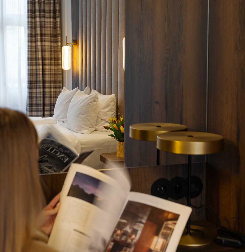 Pokój hotelowy z wysokim tapicerowanym zagłówkiem, ciepłym oświetleniem ściennym, białymi poduszkami, kratkowymi zasłonami i świeżymi kwiatami