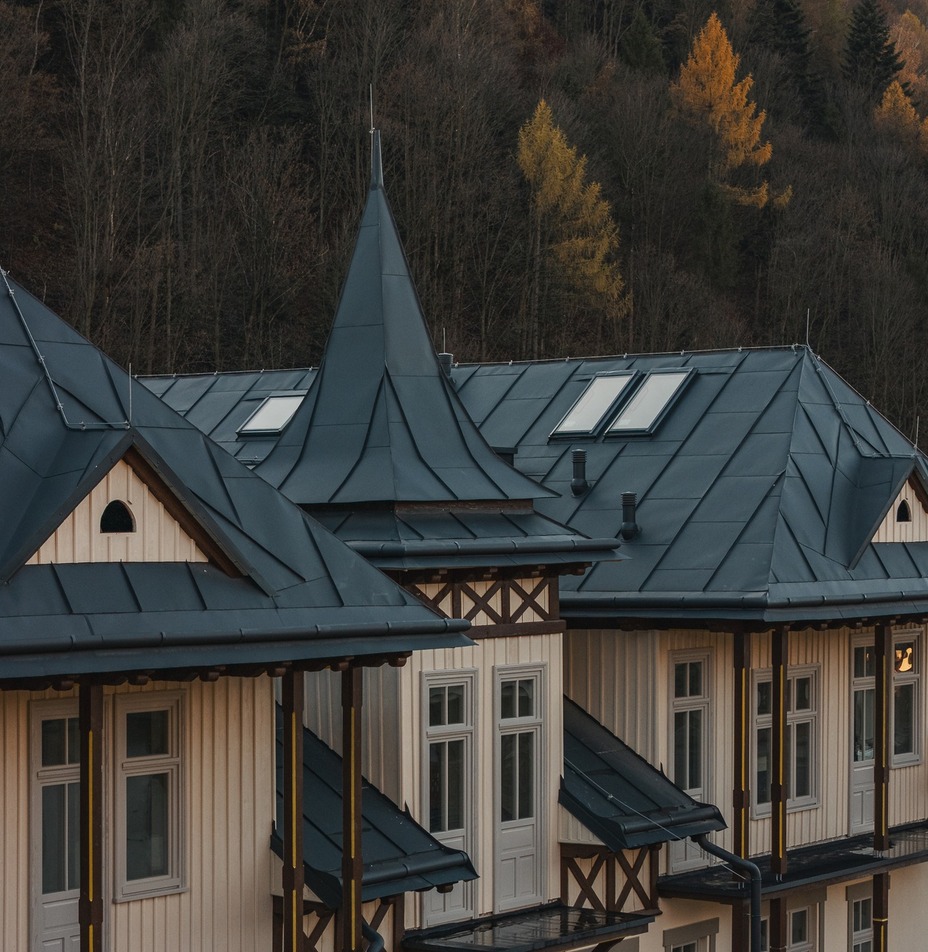 Górski hotel z ciemnym metalowym dachem i wieżyczką, oknami dachowymi i drewnianymi balkonami nad jesiennym lasem.