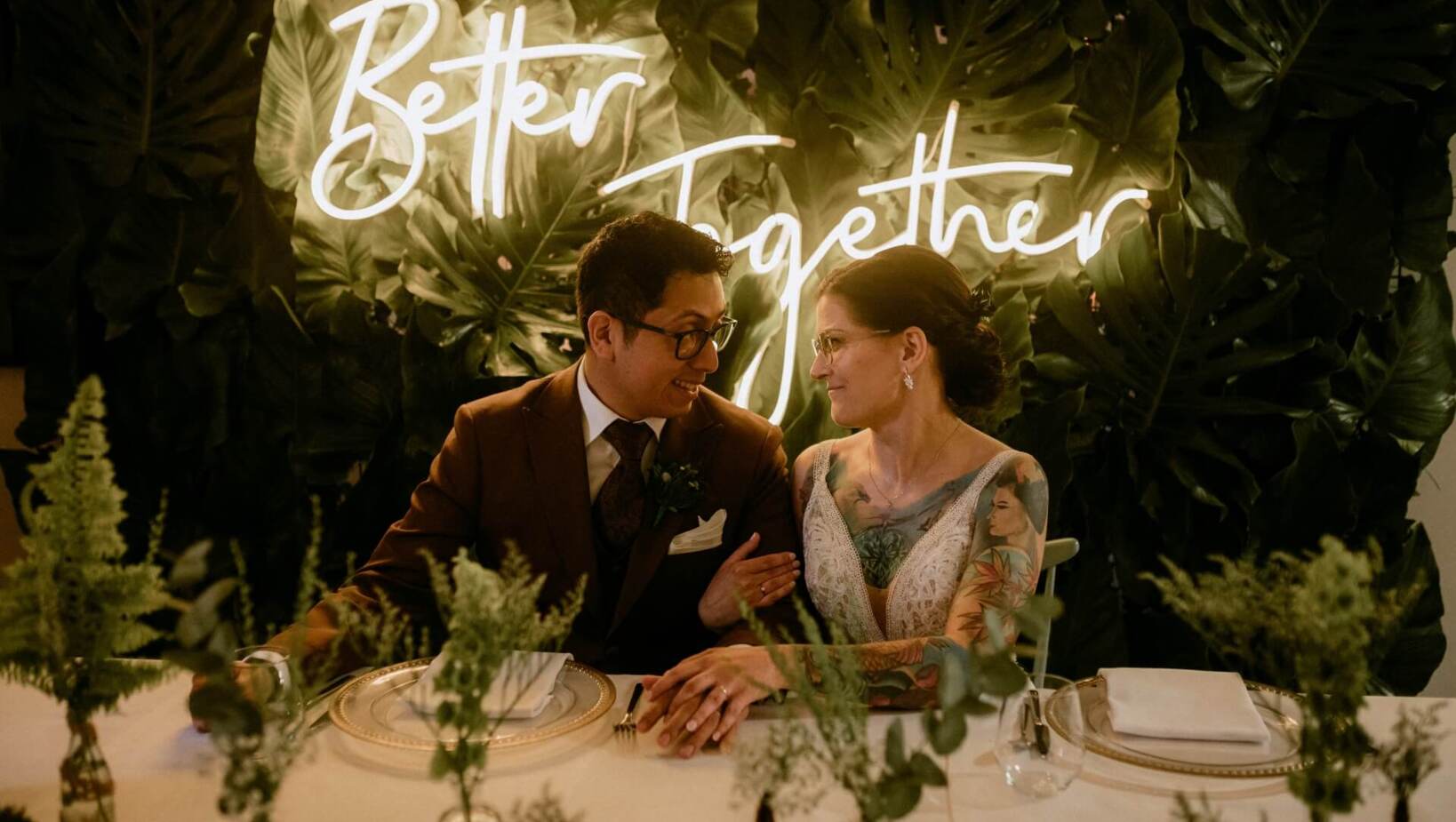 Bride and groom at an elegantly set table with a neon sign in the background